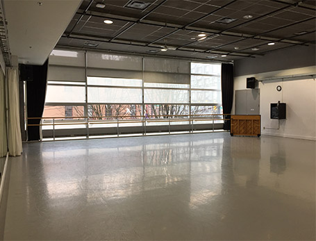 An empty dance studio with an upright piano. It has mirrors, windows with half drawn curtains and barres against the windows. The floor is shiny and reflective in the sun