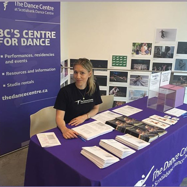 Volunteer sits at a desk filled with Dance Centre brochures and information in the lobby of Scotiabank Dance Centre