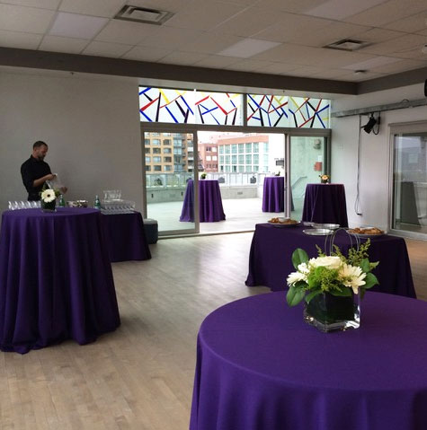 A bright dance studio set up for an event with tables covered in purple tablecloths and flowers on them. The doors to the patio are open and there are two more tables outside.
