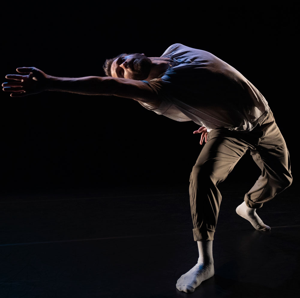 A dance artist leans over with an arm stretched out laterally. He is in a slight lunge and is wearing street clothes and white socks.