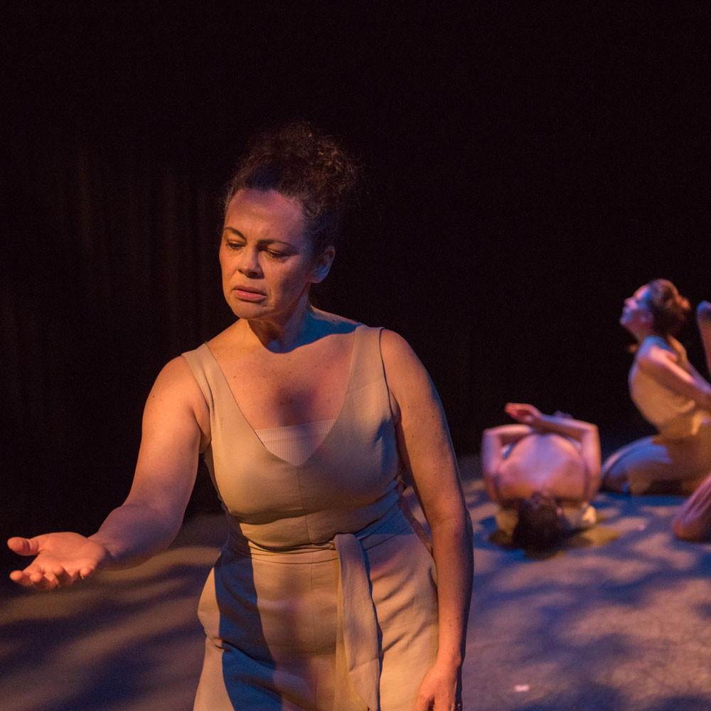 Dance artist from Raven Spirit Dance Society performs in a black box theatre. She has one arm outstretched. Behind her other dance artists pose on the floor.