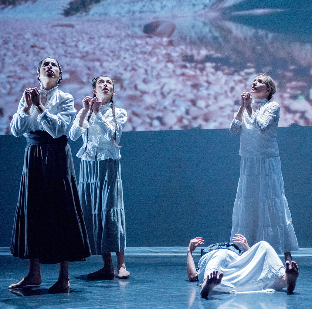 Three dance artists stand on stage with their hands clasped together. They are all wearing long skirts and long shirts. There is a fourth person lying on the floor.