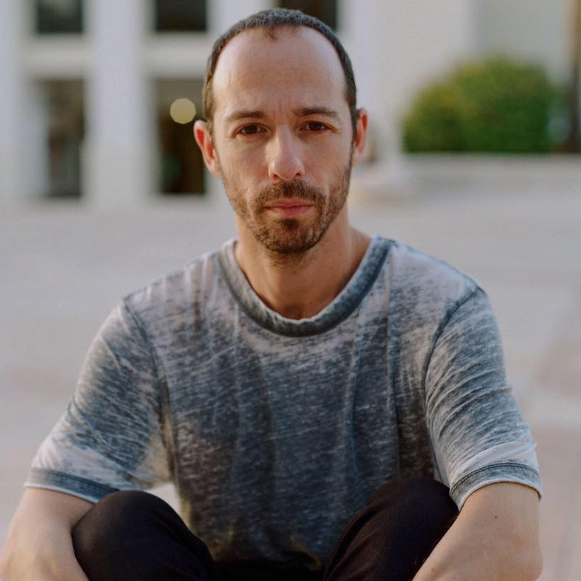 Dance Artist Hillel Kogan sitting outside. He is wearing a blue grey shirt and black pants.