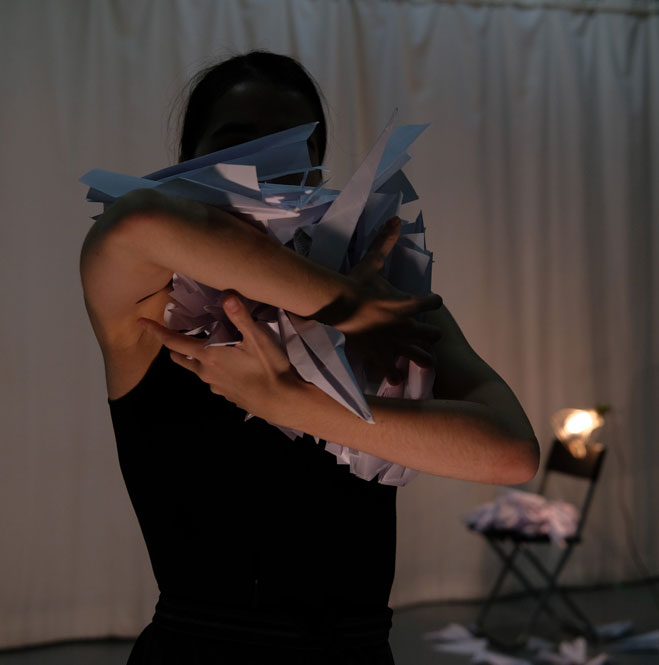 Dance artist Daria Mikhaylyuk poses mid performance. They are holding numerous paper air planes in their arms and they are obscuring the dancers face.