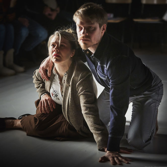 Dance artists Maria Nurmela and Ville Oinonen crouch over on stage with their arms around each other, mid performance
