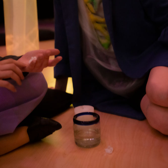 A glass half full of clear liquid sits on a wood floor directly in front of a white candle, unlit. Two people sitting are behind the items, with just their lower bodies and legs in the photograph.