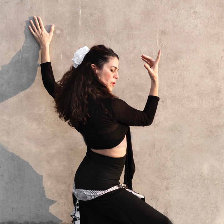 Liat Har Lev poses against a tan wall on a sunny day. She is wearing a black traditional flamenco outfit and has a large white flower in her hair.