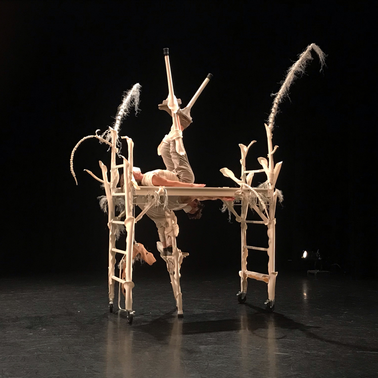 A white structure stands in the middle of a black stage. It has ladders as it's legs and a plank across the top. There are long white sticks and feathers sticking out the top. Two dance artists interact with the structure - one hanging from the plank with one leg on the ground and the other lying on the top of the plank with legs in the air. Both dance artists are wearing white and have stilts on.