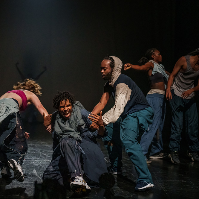 A group of artists move on stage. One is being pulled across on the stage by two others. They are wearing baggy clothes, some of which that are torn. There is smoke behind them.