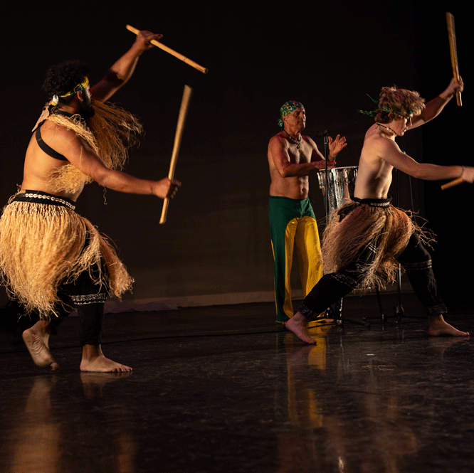 Two dance artists mid performance. They are wearing black pants and grass skirts and are holding long wooden sticks up in the air
