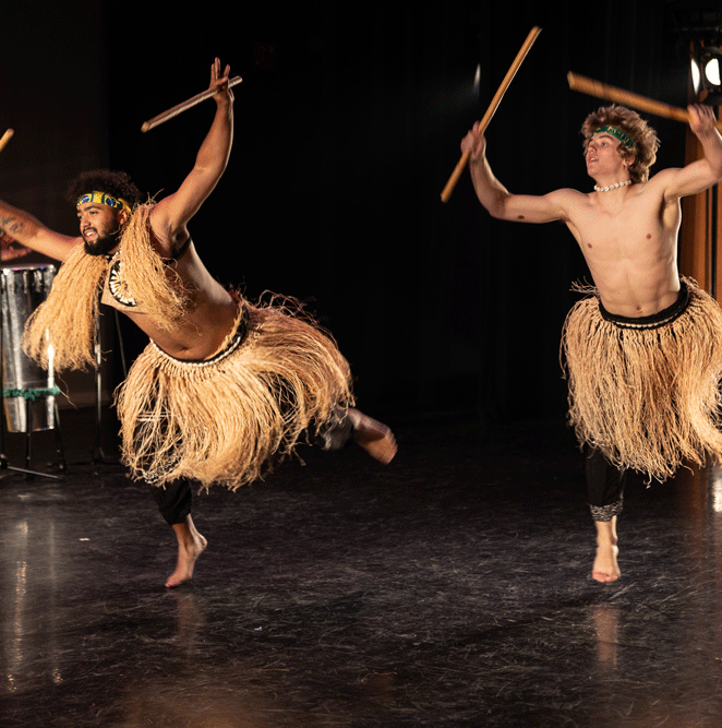 Two dance artists mid performance. They are wearing black pants and grass skirts and are holding long wooden sticks up in the air