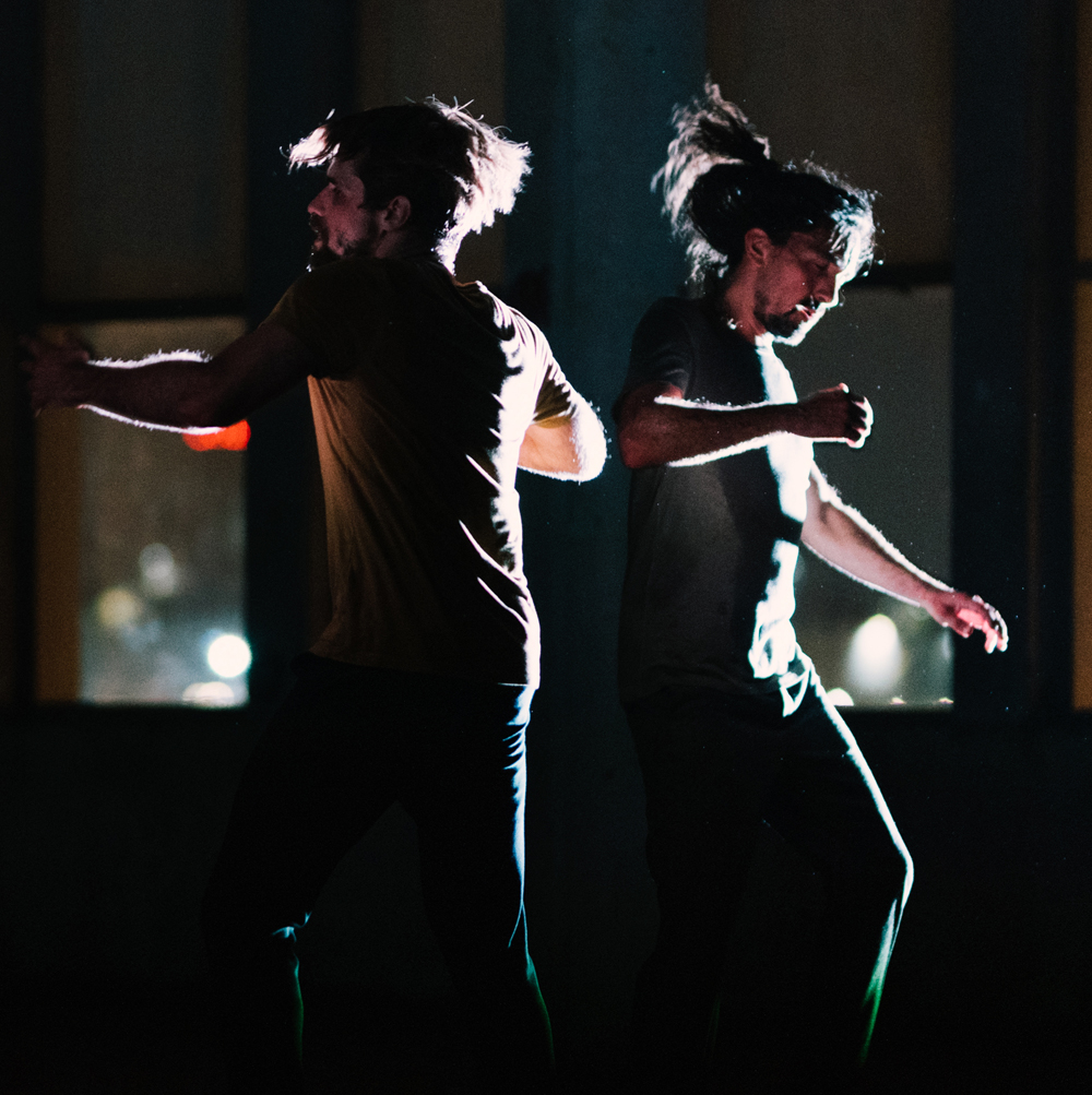 Two dance artists move and twirl on a dark stage with only a small amount of light illuminating them. Their hair is in motion as they move and there is a small window behind them.