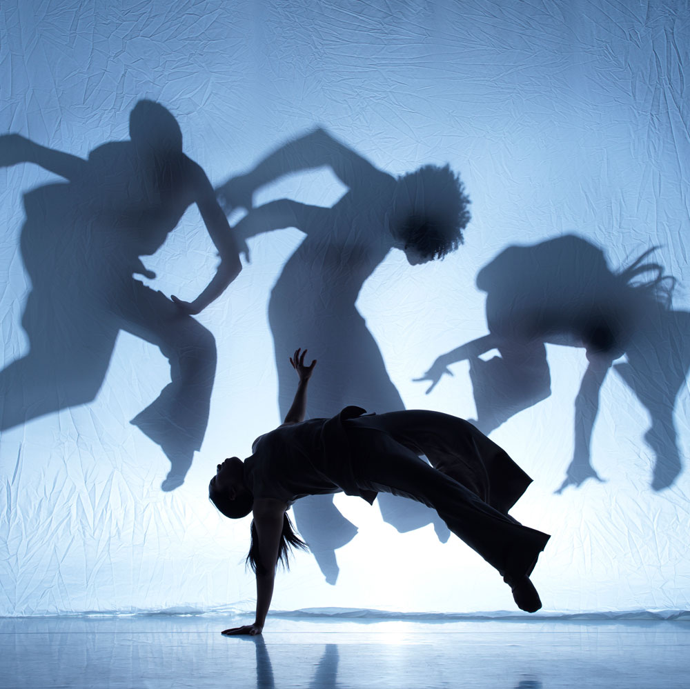 A dance artist balance on one arm mid flip. They are back lit to look like the silhouette's of other dance artists projecting on the screen behind them.