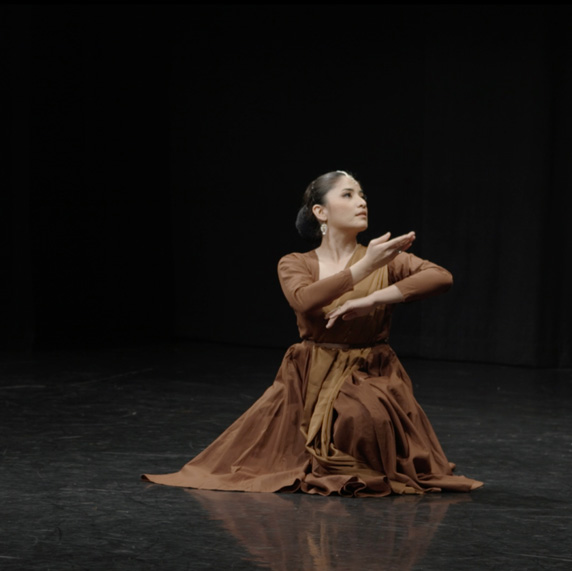A dance artist sits on a dark stage. She is wearing a brown dress and Kathak jewelry and her arms are bent up in front of her and she is looking to her left.