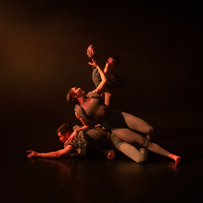 Three dance artists pose on a dark stage. They are wearing renaissance type shrugs and are lying on top of each other.
