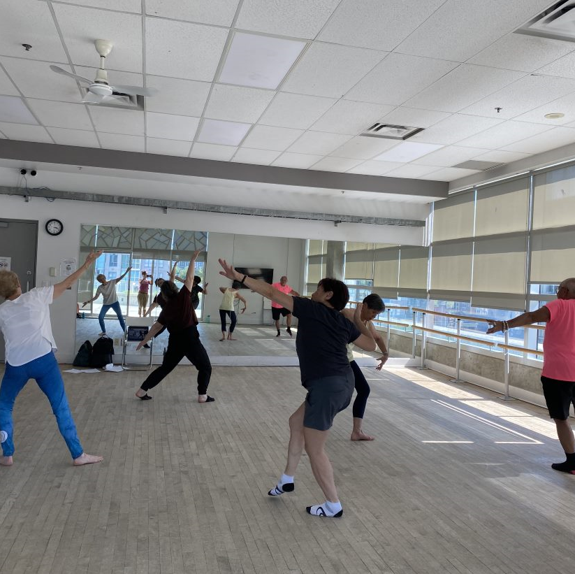 A group of seniors dancing in a bright studio. They are all wearing workout clothing and are moving around the studio with their arms extended.