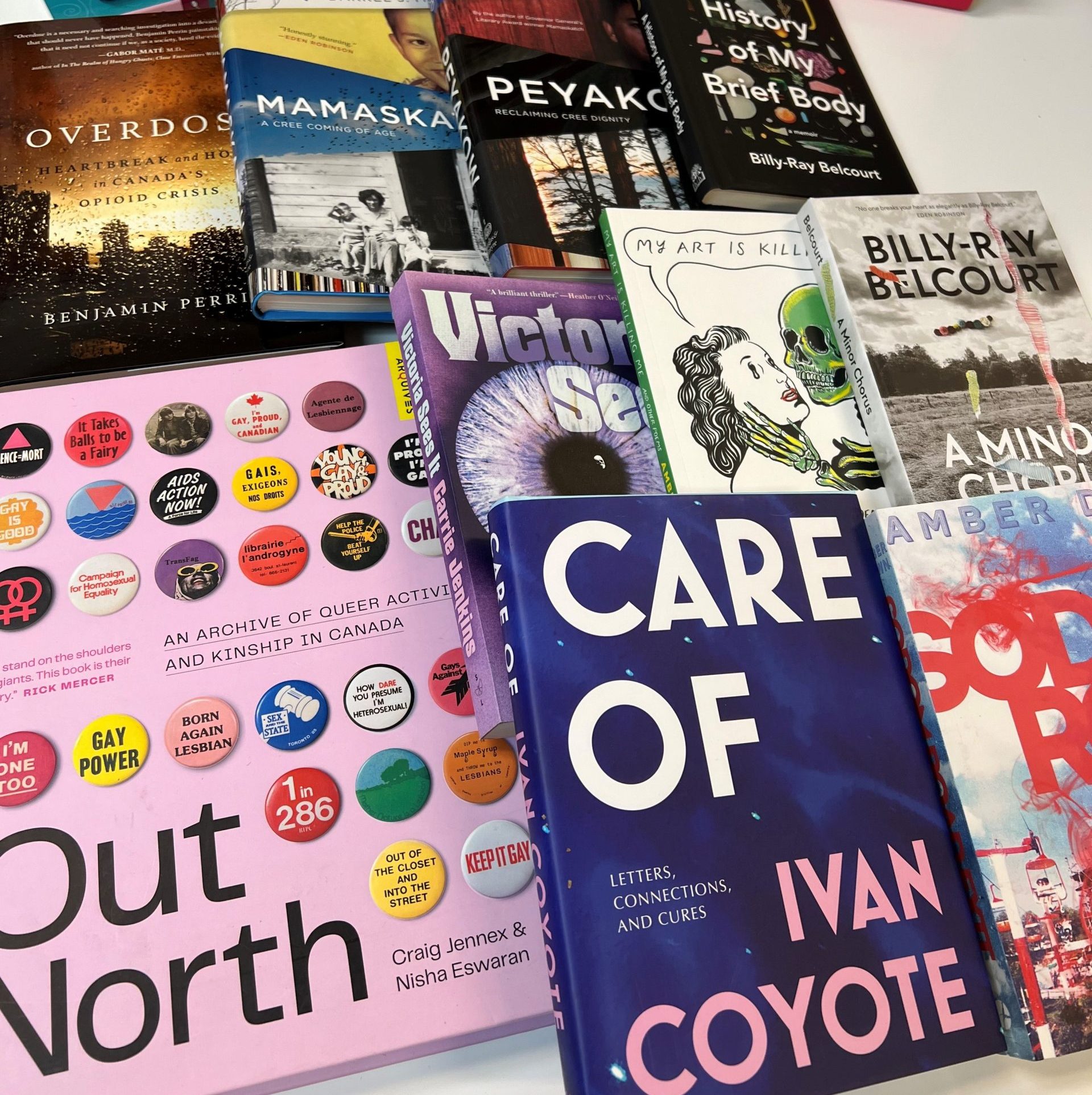 A variety of books, some hard cover, some paper back, spread out on a table side by side, some sitting on top of others.