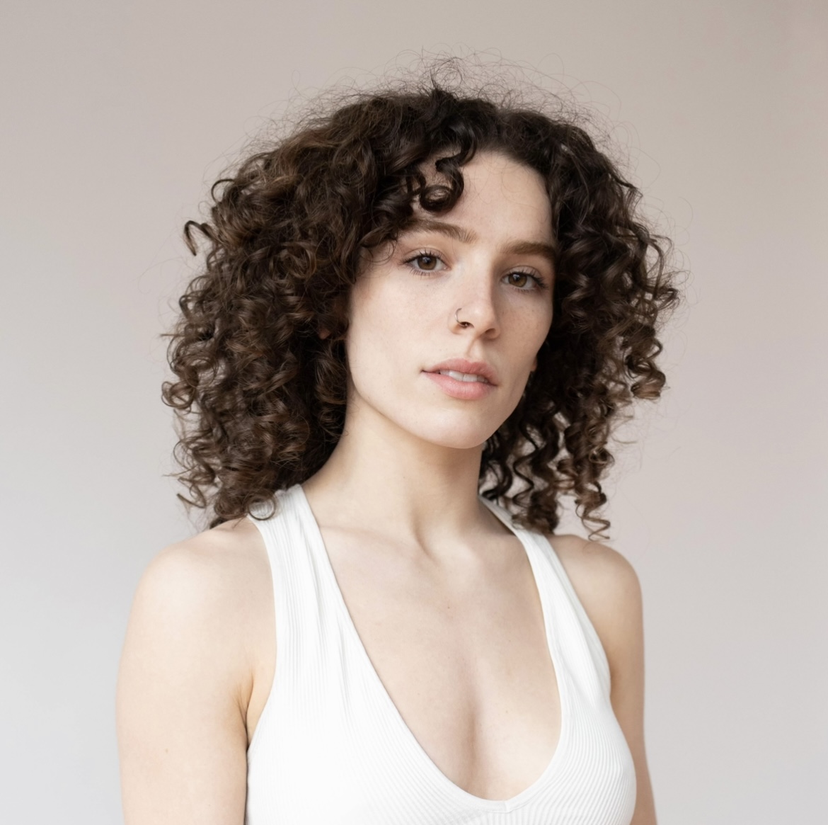 Headshot of Brenna Metzmeier. She has brown curly hair and a white tank top and is standing in front of a peach background.