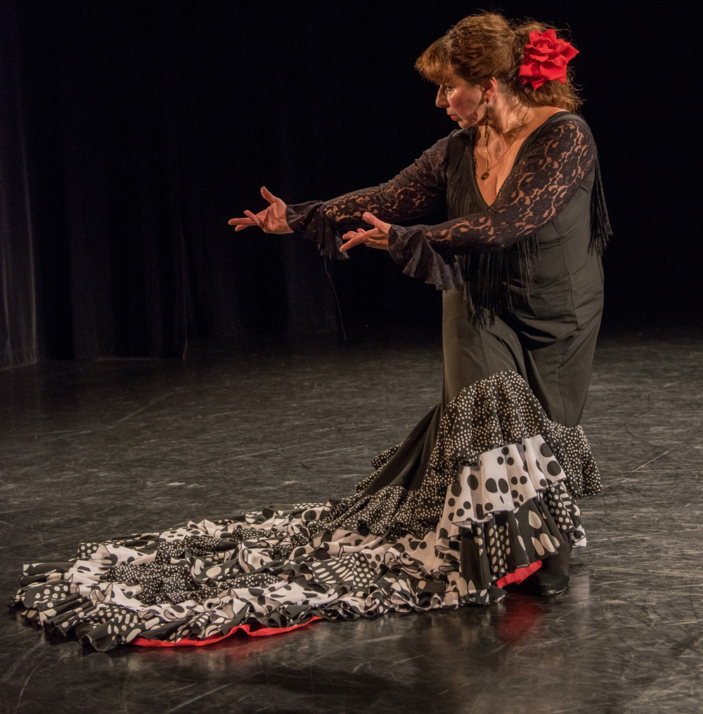 Linda Hayes poses on a dark stage. She is wearing a long black and white traditional flamenco dress with a red flower in her hair.