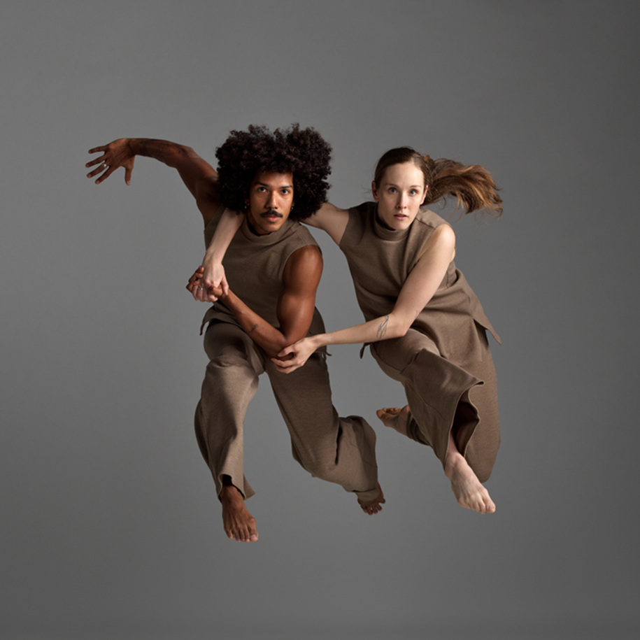 Two dance artists jump together. They are holding hands with one artist's arm draped over the others neck. They are both wearing tan brown and the back ground is also brown grey.