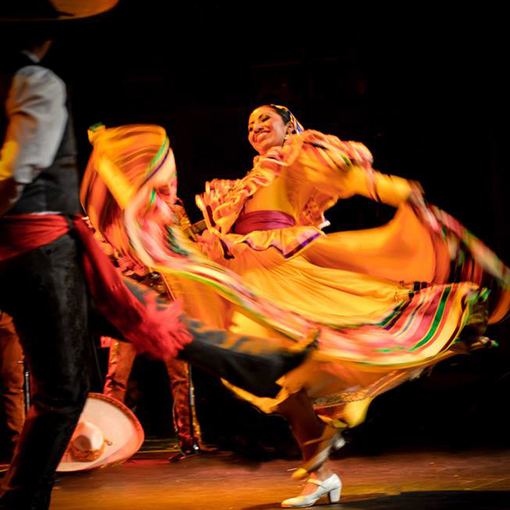 A dance artist smiles mid performance. She is wearing a bright traditional orange dress with frills and multi-coloured trim. She is twirling the skirt and you can see her white shoes underneath.