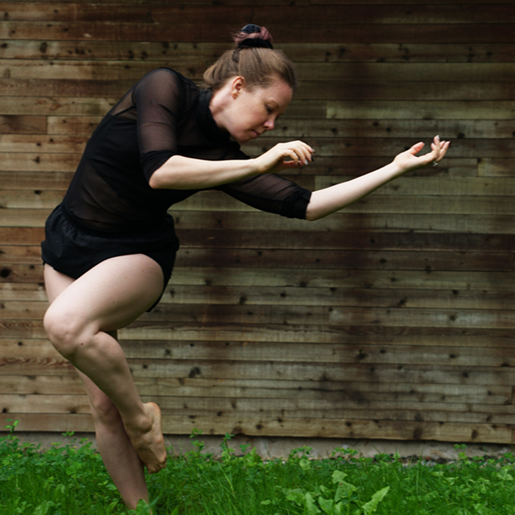 Alina Sotskova dances on grass in front of a wood wall. She is wearing a long sleeve black leotard and is bending sideways with her arms extended and one leg bent up.