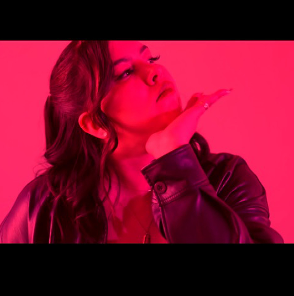 A screenshot of Raven Grenier. She is looking away from the camera with her chin in her hand. She is wearing a black jacket and the whole image is tinted red.