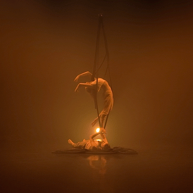 Two dancers on a hazy orange stage. One is hanging from ropes and the other is lying on the stage, holding the ropes.