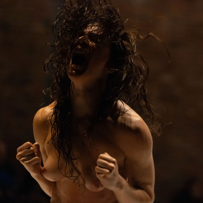 A dance artist mid scream. Their dark long hair obscures their face. They are topless.