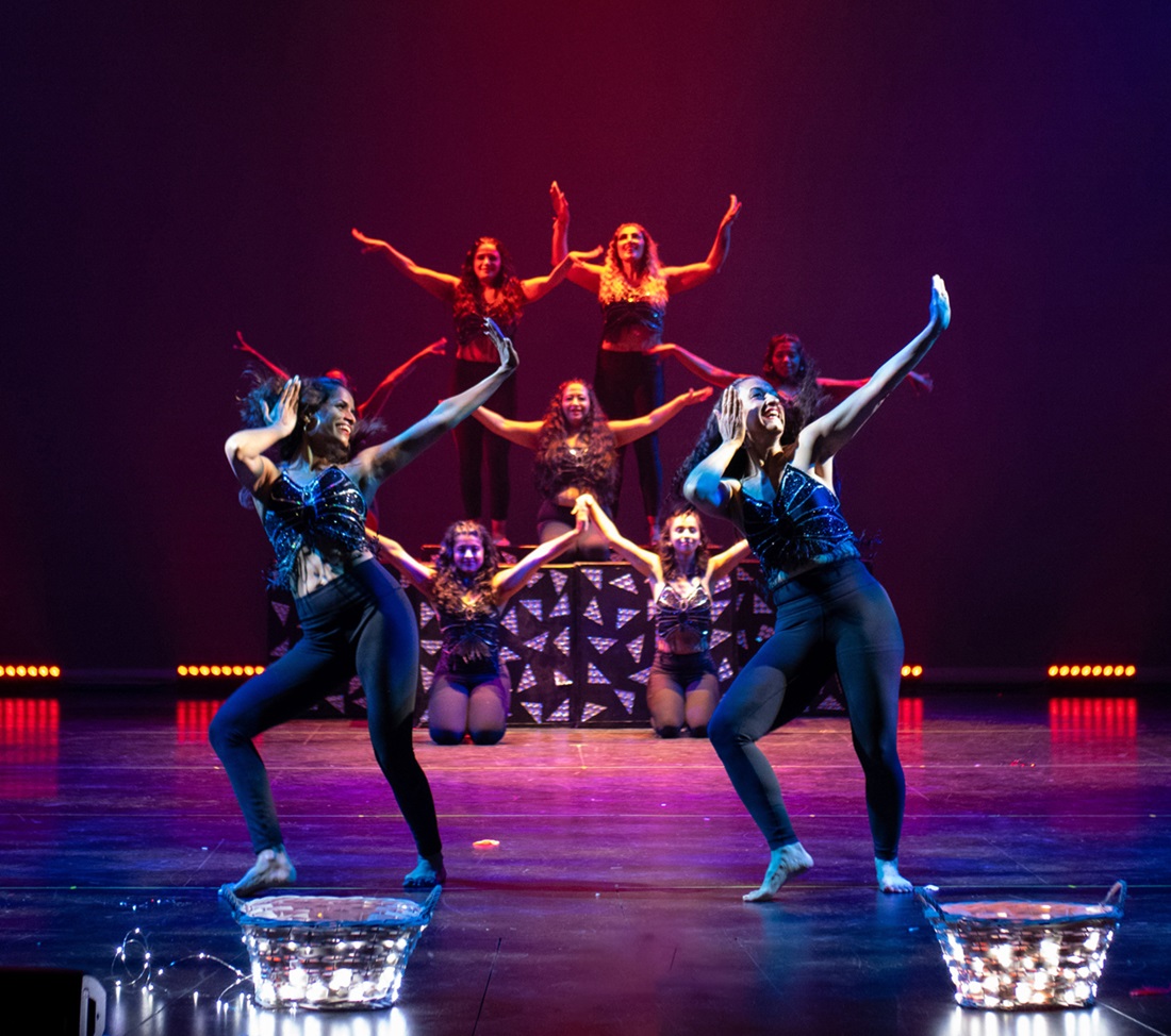 A group of dancers on stage mid performance. There are two at the front and a number at the back arranged around some boxes. They all have their arms raised in various formations.