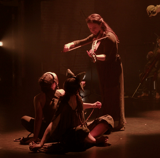 Two dance artists sit on a stage looking up at a violinist playing above them. The dancers are sitting back to back and you can see that they are wearing animal masks.