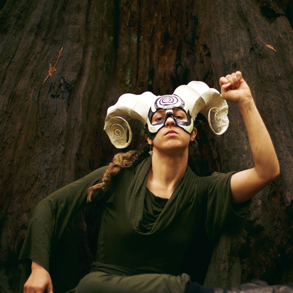 A dance artist poses in the forest. She is wearing a green tunic and a mask in the form of a ram with long curly horns. Their hair is in a long braid and she has one arm raised up in a fist.