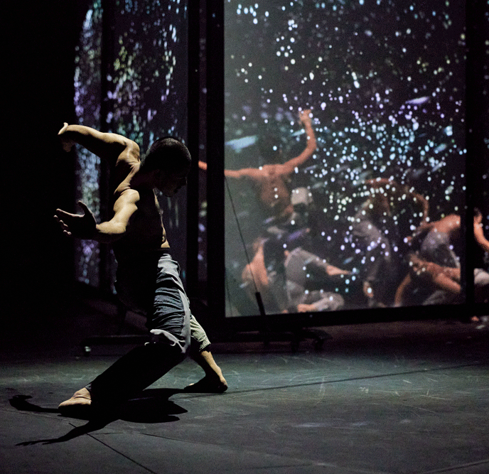 A dance artist stands with his legs and arms extended. He is wearing only blue pants and behind him is a glass wall reflecting colourful lights and other dancers in similar poses and outfits.