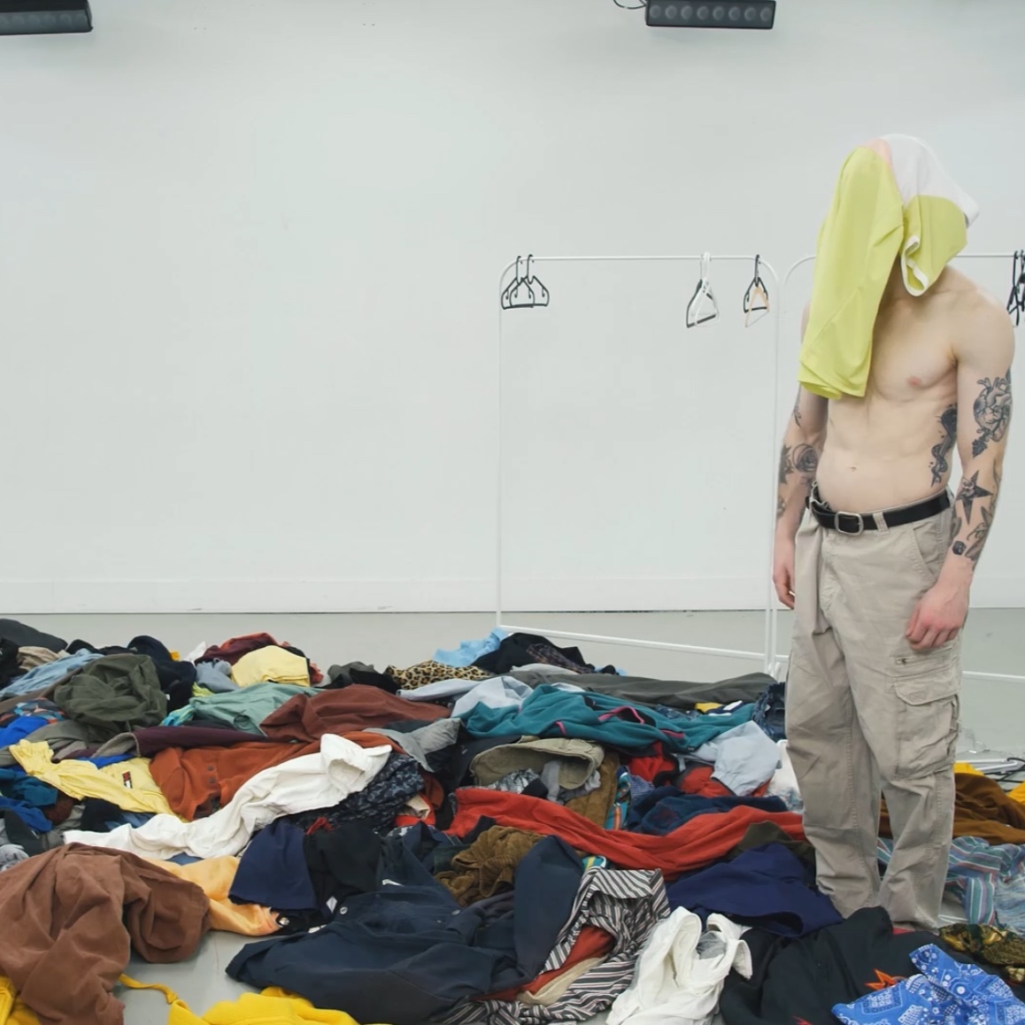 A dance artist in a bright studio. They have on khakis and tattoos up their arms and chest. There is a shirt over their face and the floor is covered in clothes.
