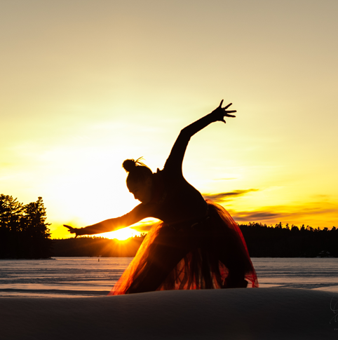 A dance artist poses outside. They are backlit by the setting sun so you can only see the silhouette of the dancer who is bending over, arms extended and is wearing a long skirt.